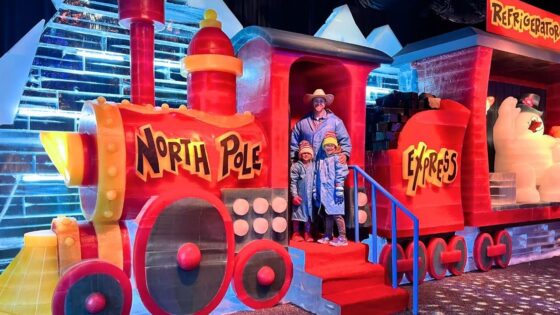 Colorful ice sculpture train labeled 'North Pole Express' with a man and two children posing on red steps in winter coats.