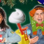 Two smiling hosts in denim jackets pose with holiday props: a wrapped gift on the left and a cartoon monkey-costume cutout on the right, against a Christmas backdrop with greenery and ornaments.