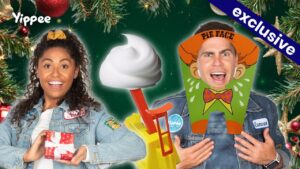 Two smiling hosts in denim jackets pose with holiday props: a wrapped gift on the left and a cartoon monkey-costume cutout on the right, against a Christmas backdrop with greenery and ornaments.