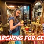 Young woman with glasses holds a small gold goblet in a treasure-filled jewelry shop, surrounded by gold coins, necklaces, and shelves of jewelry.