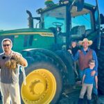 A group of people posing with a large green John Deere tractor with yellow wheels on a sunny farm field; harvest items in their hands suggest a harvest moment.