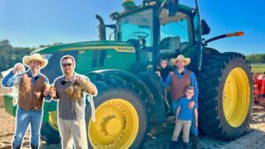 A group of people posing with a large green John Deere tractor with yellow wheels on a sunny farm field; harvest items in their hands suggest a harvest moment.