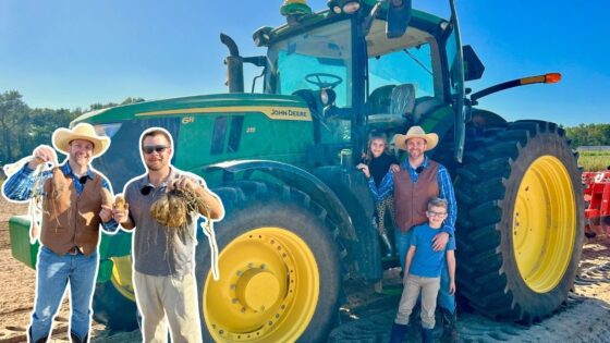 A group of people posing with a large green John Deere tractor with yellow wheels on a sunny farm field; harvest items in their hands suggest a harvest moment.