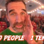 Bearded man in a green shirt in a festive tent with string lights and people in Christmas sweaters; overlay text reads '50 PEOPLE 1 TENT'