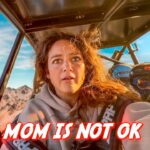 In-flight selfie of a woman with a rocky mountain landscape outside the cockpit; bold red text across the bottom reads 'MOM IS NOT OK'.