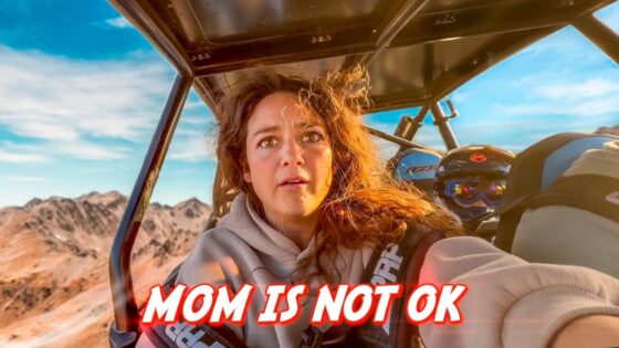 In-flight selfie of a woman with a rocky mountain landscape outside the cockpit; bold red text across the bottom reads 'MOM IS NOT OK'.