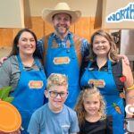 Group photo of four people in a kitchen, all wearing blue aprons with a sweet potato logo, smiling at the camera.