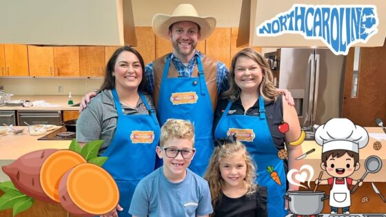 Group photo of four people in a kitchen, all wearing blue aprons with a sweet potato logo, smiling at the camera.