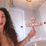 Woman with long curly hair smiling at the camera indoors, pointing to a bold 'Do It Yourself!' graphic on the wall.