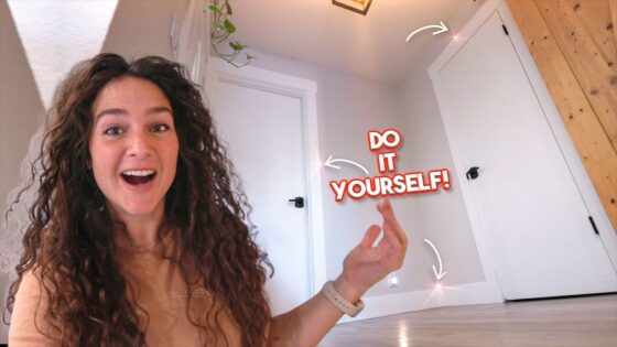Woman with long curly hair smiling at the camera indoors, pointing to a bold 'Do It Yourself!' graphic on the wall.