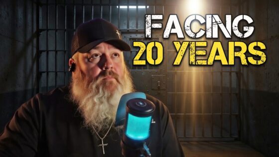 Bearded man wearing a cap speaks into a blue microphone in a dim jail-cell backdrop, with large yellow and white text reading 'FACING 20 YEARS' in the foreground.
