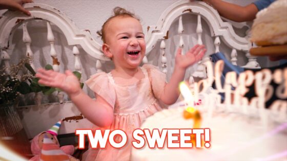 Smiling toddler in a pink dress claps at a birthday party with a cake and candles in the foreground, and a decorative white backdrop behind.