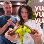 Couple in a kitchen holding a broken chocolate bar with bright green goo dripping from it, surprised expressions show curiosity. (YUMMY text overlay).