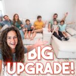 Smiling woman in the foreground taking a selfie in a bright living room, with a large white sectional and a family on the couch behind, overlaid with bold 'BIG UPGRADE!' text.