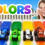 A cheerful girl sits beside rainbow-colored paint trays with six toy trucks in rainbow order, beneath a big 'COLORS' sign.