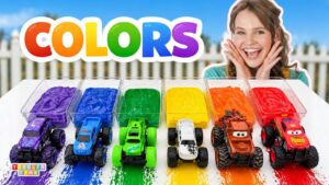 A cheerful girl sits beside rainbow-colored paint trays with six toy trucks in rainbow order, beneath a big 'COLORS' sign.