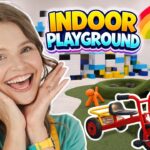 Cheerful woman in foreground with a colorful indoor playground and toy structures behind her.