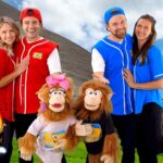 Four performers in red and blue uniforms pose with two monkey mascot characters outdoors, next to a colorful 'Good News Guys' sign and a smiling sun in the sky, conveying a family-friendly entertainment vibe.