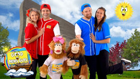 Four performers in red and blue uniforms pose with two monkey mascot characters outdoors, next to a colorful 'Good News Guys' sign and a smiling sun in the sky, conveying a family-friendly entertainment vibe.