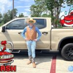 Person in a tan vest, blue shirt, jeans, and cowboy boots stands beside a beige pickup truck at a car wash, with cartoon bubbles and a red cartoon car in the foreground.