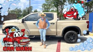 Person in a tan vest, blue shirt, jeans, and cowboy boots stands beside a beige pickup truck at a car wash, with cartoon bubbles and a red cartoon car in the foreground.