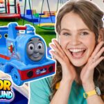 Joyful woman with hands on her cheeks beside a blue Thomas the Tank Engine ride-on train in an indoor playground.