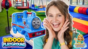 Joyful woman with hands on her cheeks beside a blue Thomas the Tank Engine ride-on train in an indoor playground.
