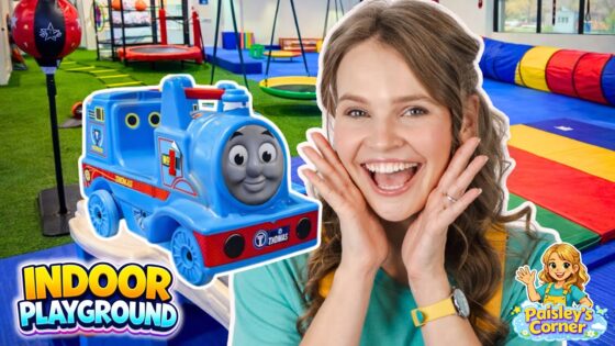 Joyful woman with hands on her cheeks beside a blue Thomas the Tank Engine ride-on train in an indoor playground.