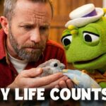 Bearded man holding a small chick wrapped in a blanket, while a green puppet gazes on; bold 'EVERY LIFE COUNTS' text overlays with Iggy and Mr. Kirk branding and Brave+ logo.