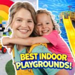 Smiling woman hugs a cheerful girl in a colorful indoor playground with a rainbow, blocks, tricycle and yellow slide in the background.