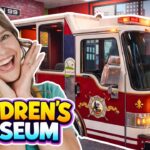 Smiling woman posing beside a red fire truck exhibit at a children's museum display, with 'Children's Museum' text overlay.