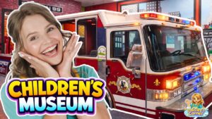 Smiling woman posing beside a red fire truck exhibit at a children's museum display, with 'Children's Museum' text overlay.